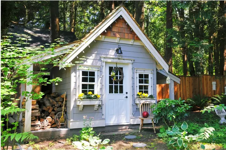 Grey garden studio with a steep gable, cedar shingle gable end, glazed door, window boxes, and an open lean-to holding stacked firewood
