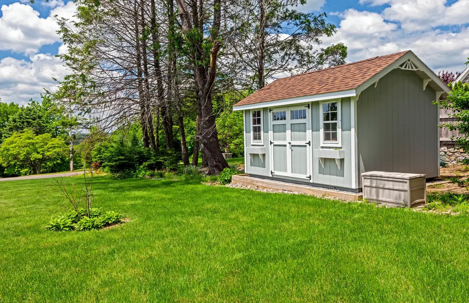 Sage green gable-roof storage shed with white double doors and windows, sitting on a gravel base