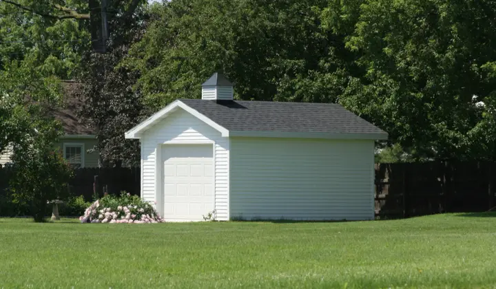 White clapboard workshop shed with a single overhead garage door and a roof cupola, set at the back of a lawn