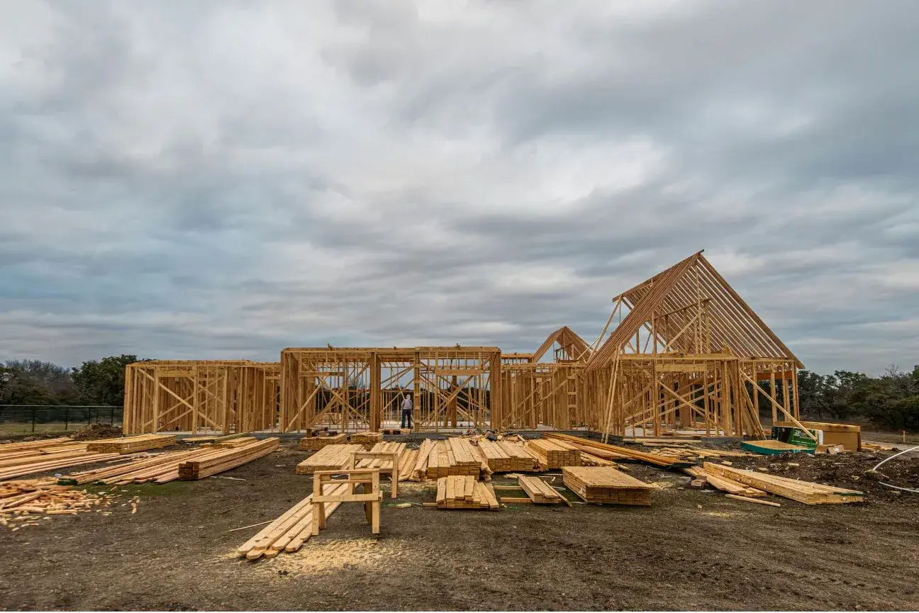 A house under construction with the exterior walls framed and the roof trusses and gable ends standing, framing lumber stacked on the ground