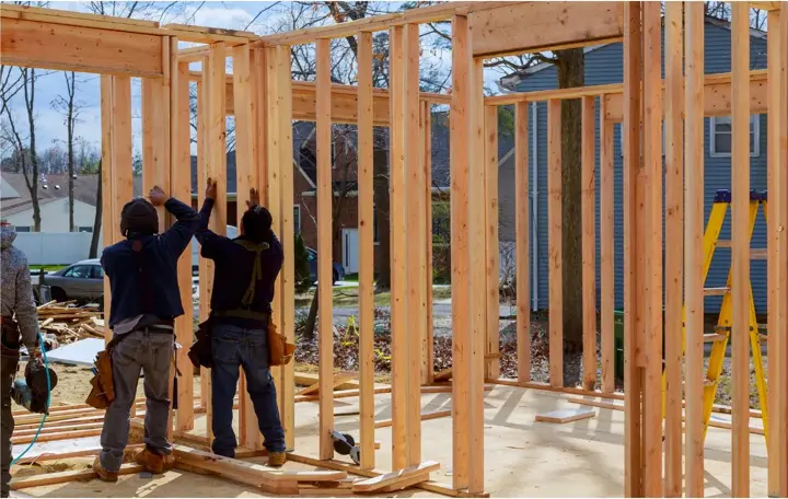 Two carpenters standing a framed exterior wall on a plywood deck beside an existing house, with headers over the openings already built in