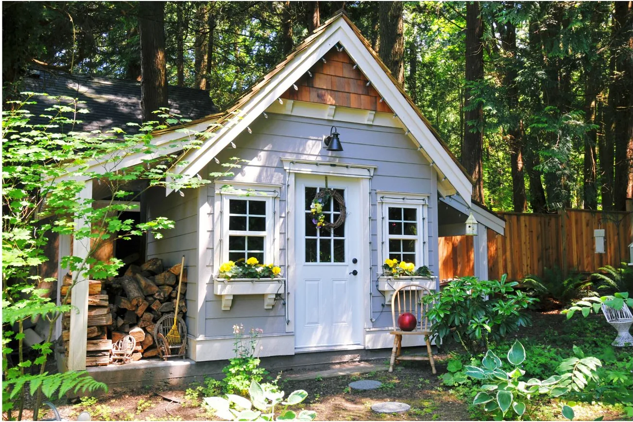 Grey garden studio with a steep gable, cedar shingle gable end, glazed door, window boxes, and an open lean-to holding stacked firewood