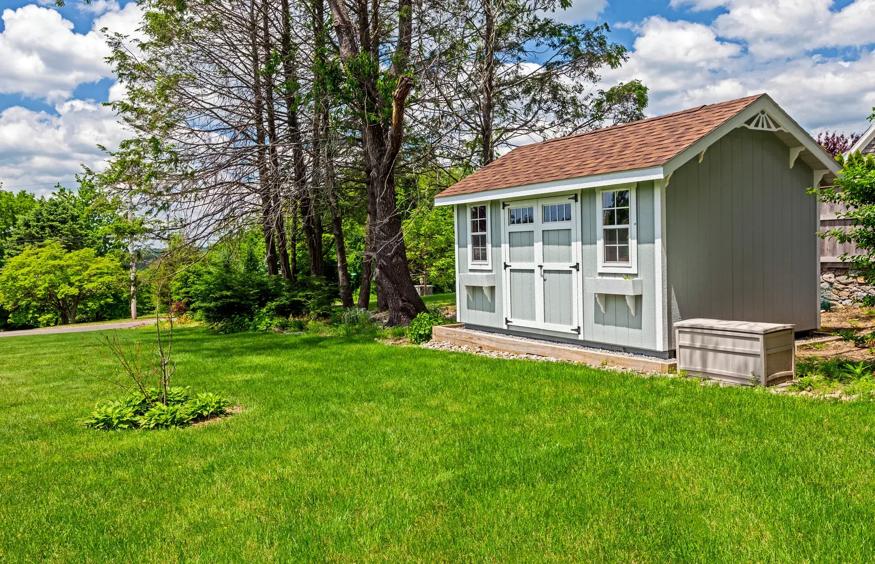Sage green gable-roof storage shed with white double doors and windows, sitting on a gravel base