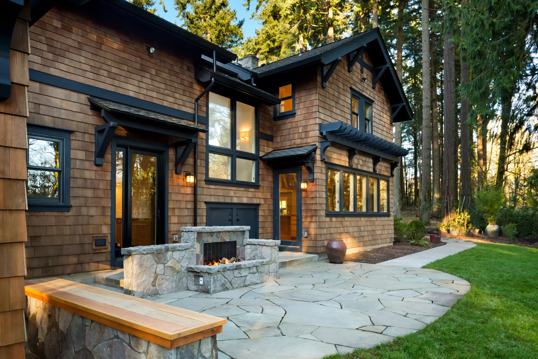 Stone outdoor fireplace built into a flagstone patio with a cedar-capped stone bench and path lighting at dusk