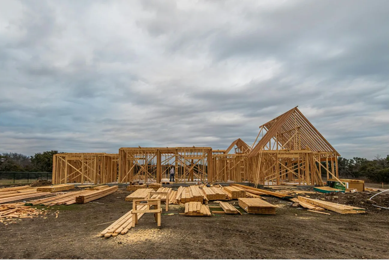 A house under construction with the exterior walls framed and the roof trusses and gable ends standing, framing lumber stacked on the ground