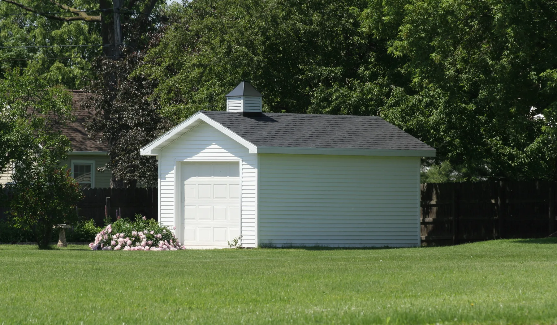 White clapboard workshop shed with a single overhead garage door and a roof cupola, set at the back of a lawn
